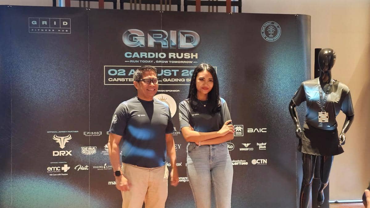 Dua orang berpose di depan backdrop acara GRID Cardio Rush 2026 di Gading Serpong dengan patung mengenakan kaus olahraga di samping mereka.