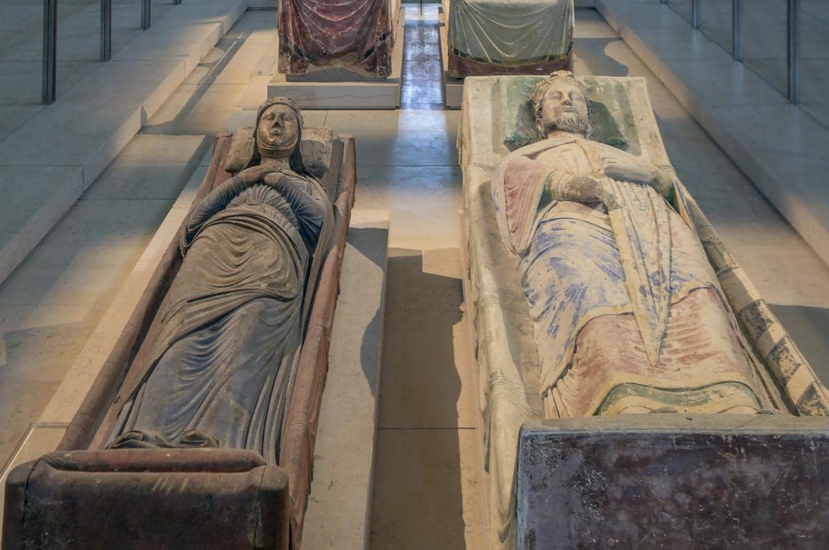 Dua sarkofagus batu berisi mumi dengan ukiran detail, menampilkan sosok berpakaian jubah panjang di ruang pameran museum.