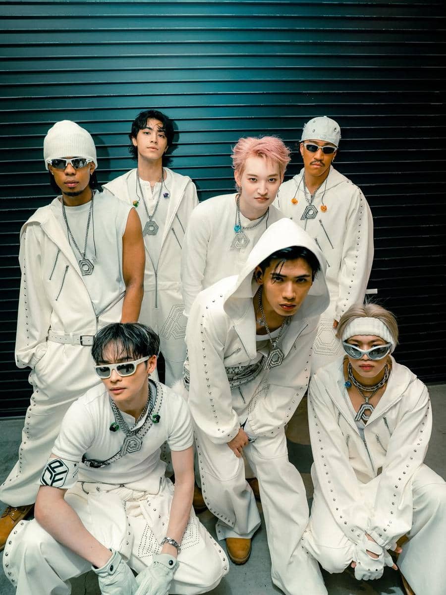 Tujuh personel boy band Jepang PSYCHIC FEVER berpose bersama mengenakan busana serba putih dengan aksesori perak di depan dinding hitam.