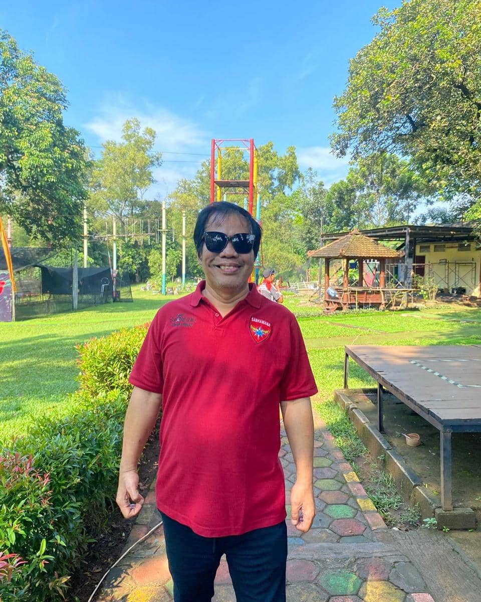 Seorang pria mengenakan kacamata hitam dan kaus polo merah berdiri di taman hijau dengan langit cerah dan pepohonan rindang.