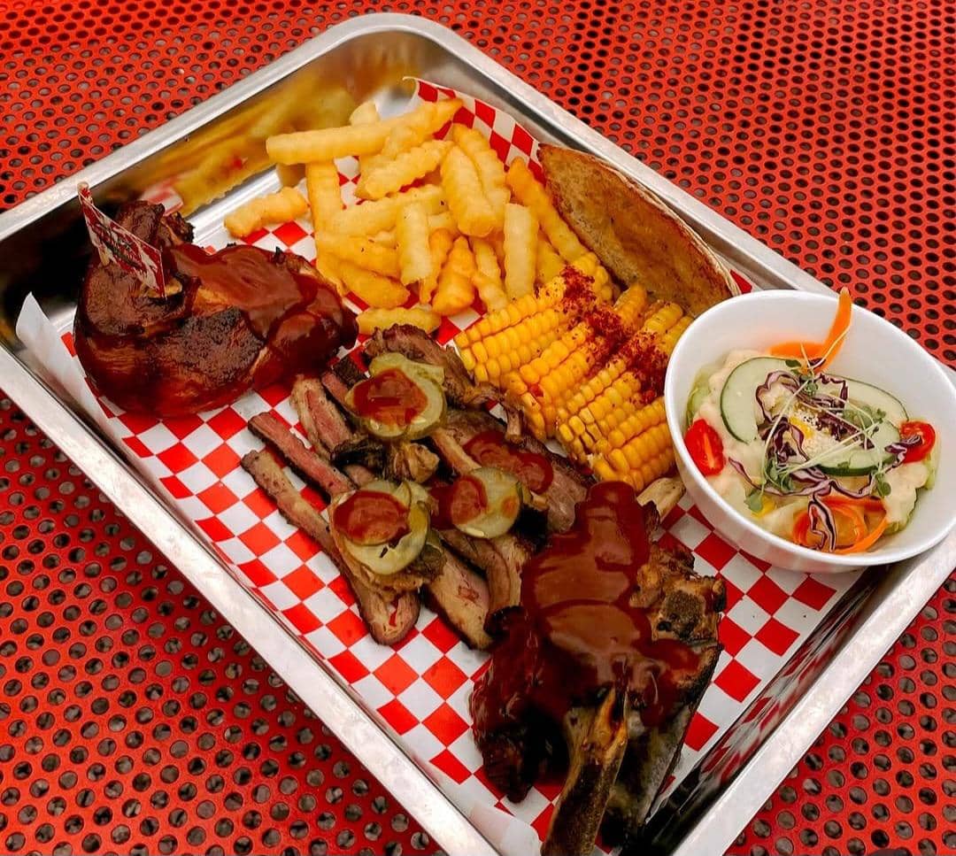Hidangan barbeque berisi iga sapi panggang dengan saus, kentang goreng, jagung manis, roti panggang, dan salad sayur segar.