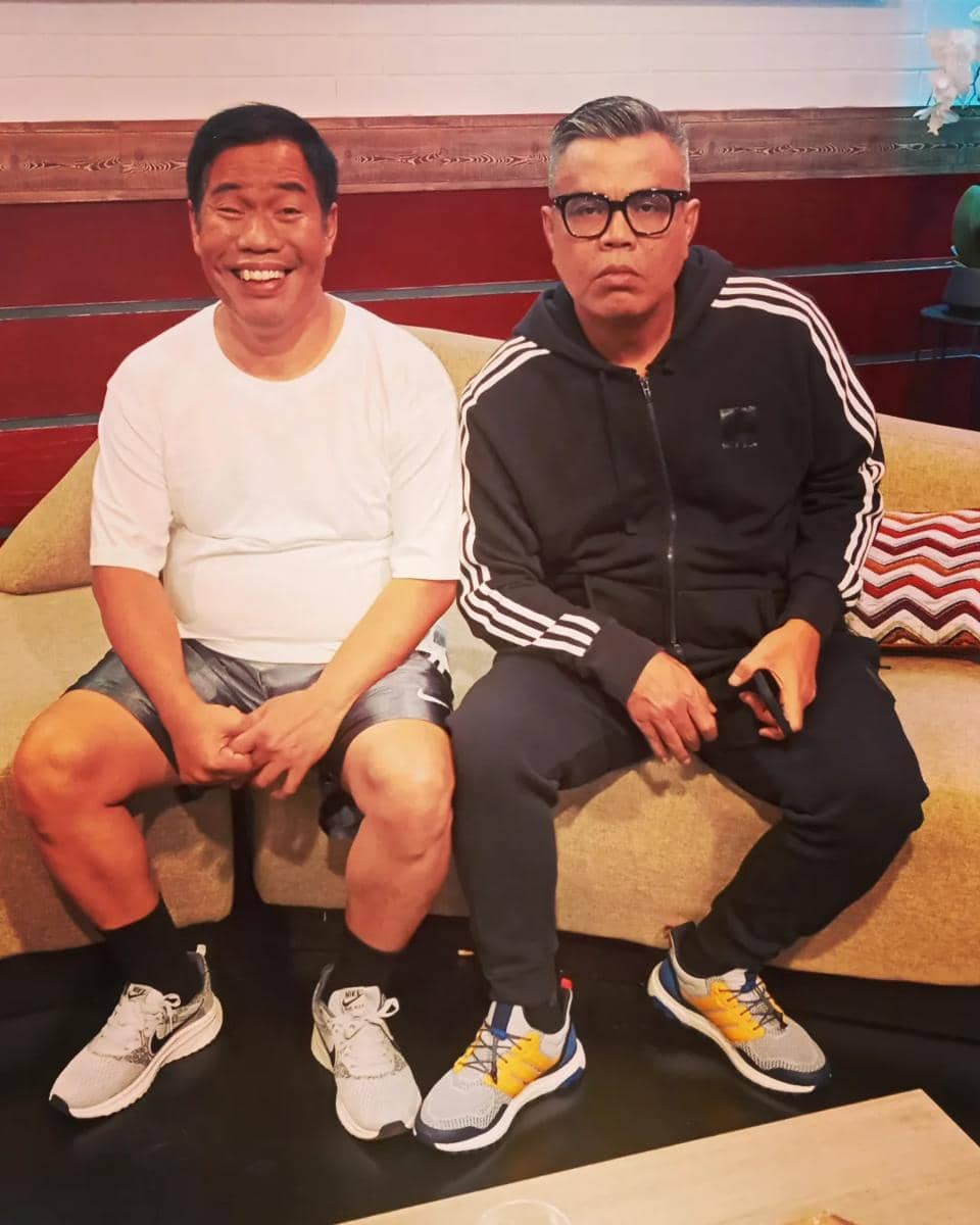 Dua pria duduk di sofa, satu mengenakan kaus putih dan celana pendek, satunya memakai jaket hitam dengan garis putih dan kacamata.