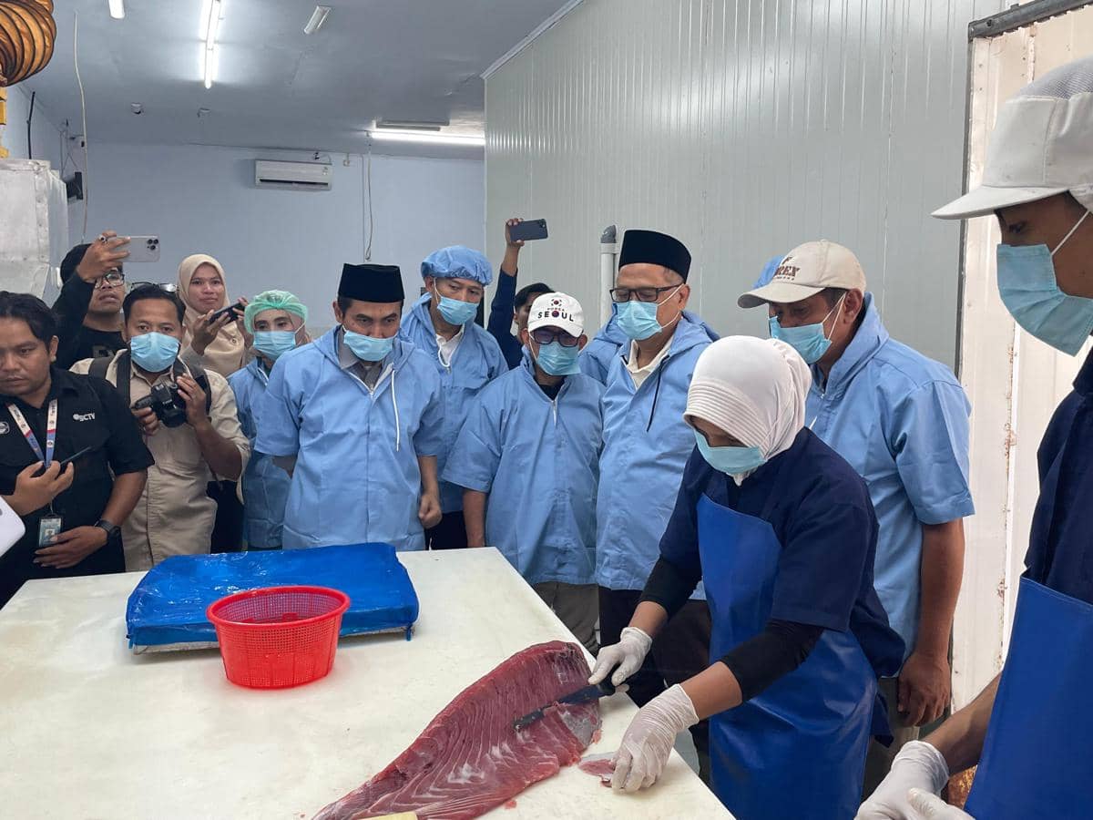 Seorang petugas memotong ikan segar di hadapan rombongan pejabat berbusana biru dan bermasker saat kunjungan kerja di fasilitas pengolahan ikan.