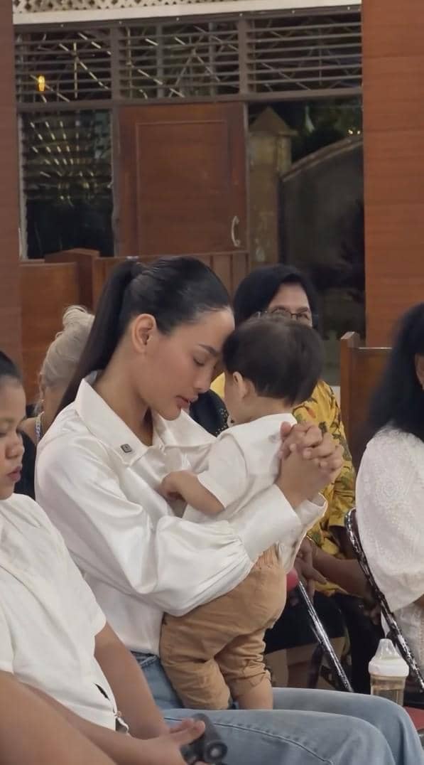 Seorang wanita mengenakan kemeja putih memeluk bayi kecil di pangkuannya sambil berdoa di dalam gereja bersama beberapa orang lain.