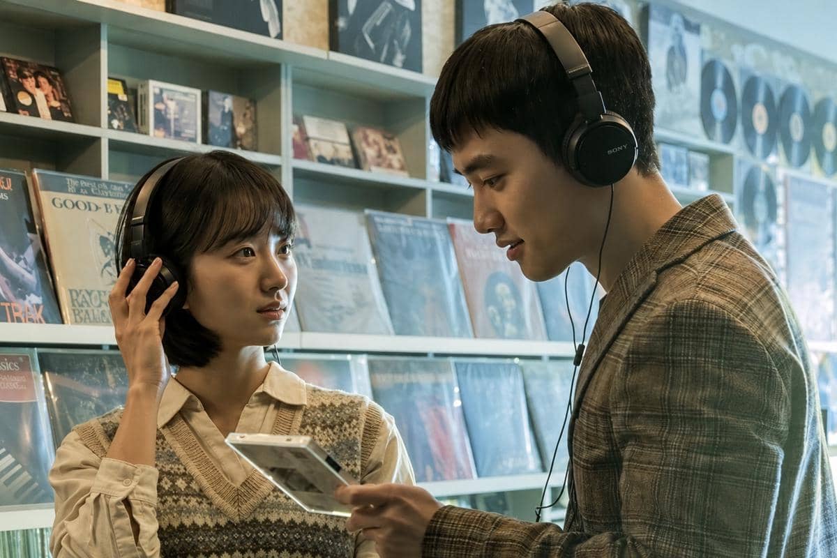 Dua karakter dalam film Secret: Untold Melody mengenakan headphone sambil memegang kaset di toko musik dengan rak piringan hitam di belakang mereka.