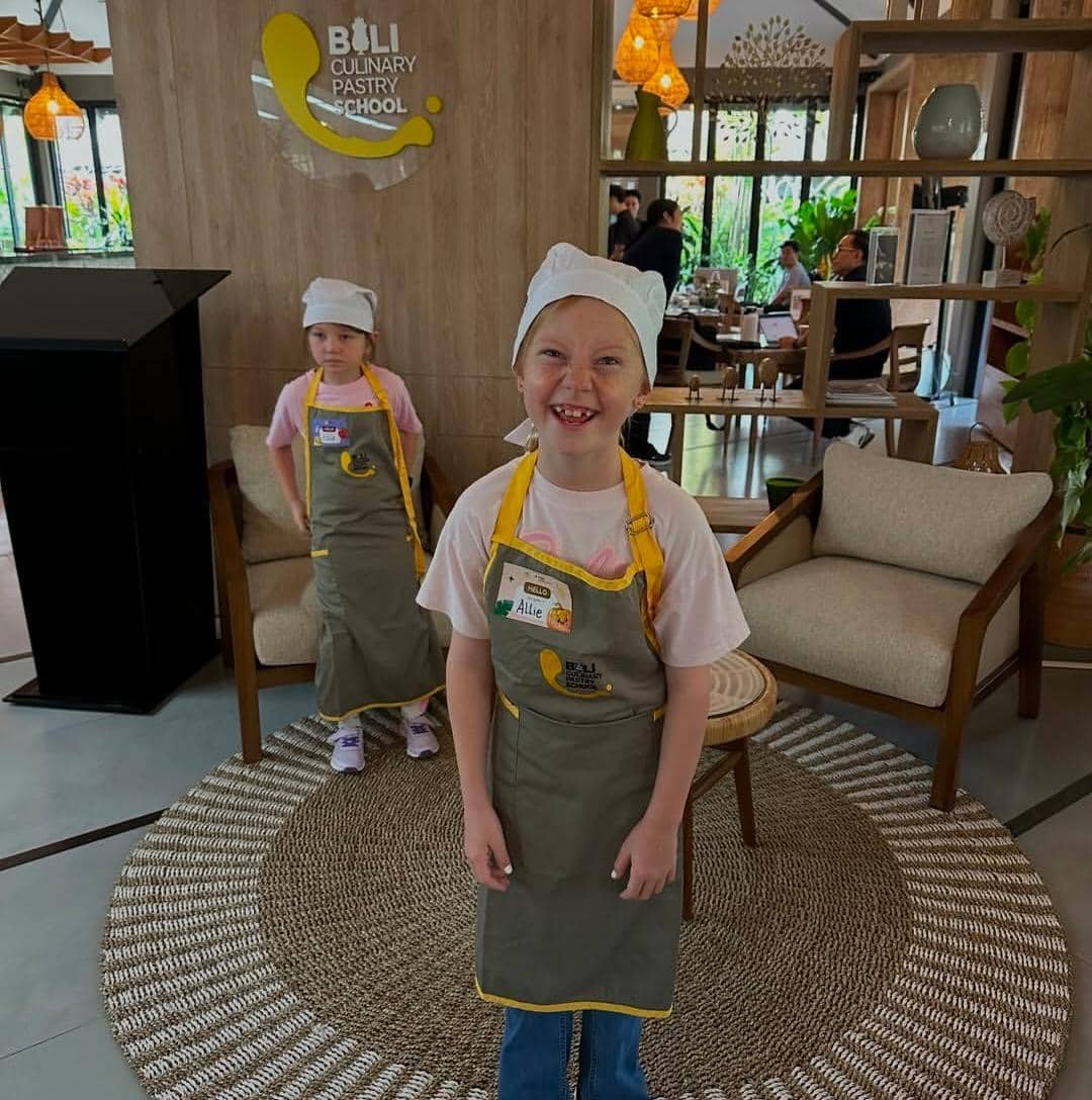 Dua anak mengenakan celemek dan topi koki kecil berpose di dalam ruang kelas memasak di Bali Culinary Pastry School.