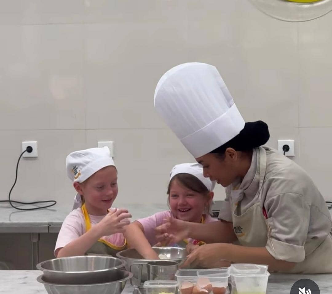 Seorang koki membantu dua anak kecil yang mengenakan celemek dan topi koki saat mengikuti kelas memasak di dapur modern di Bali.