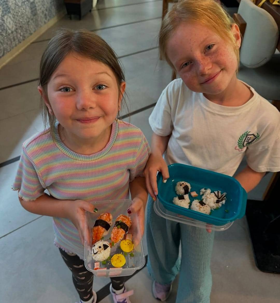 Dua anak kecil tersenyum sambil memegang kotak berisi hasil masakan berbentuk sushi dan karakter lucu di sebuah kelas memasak di Bali.