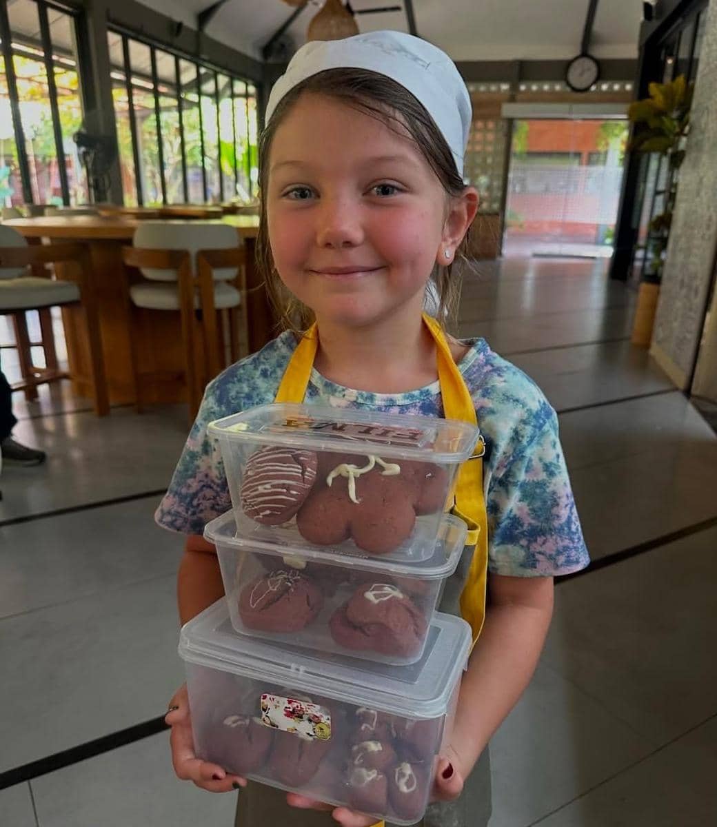 Seorang anak mengenakan celemek kuning dan topi koki memegang tiga kotak berisi kue cokelat hasil kelas memasak di Bali.