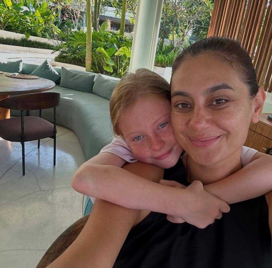 Seorang wanita tersenyum bersama anak perempuan yang memeluknya di area duduk terbuka dengan latar taman hijau di Bali.