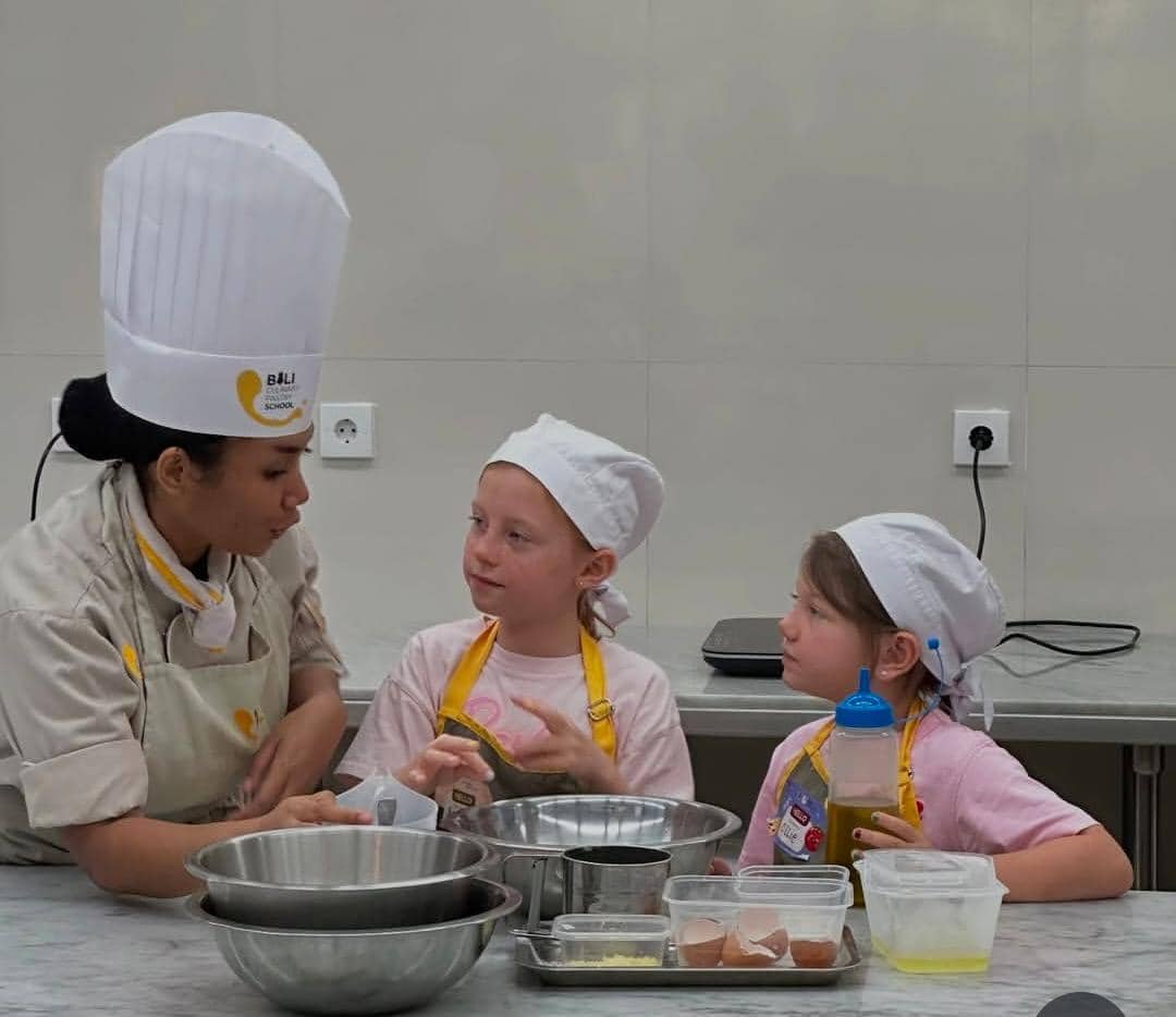 Dua anak mengenakan celemek dan topi koki belajar memasak bersama seorang instruktur di dapur sekolah memasak di Bali.