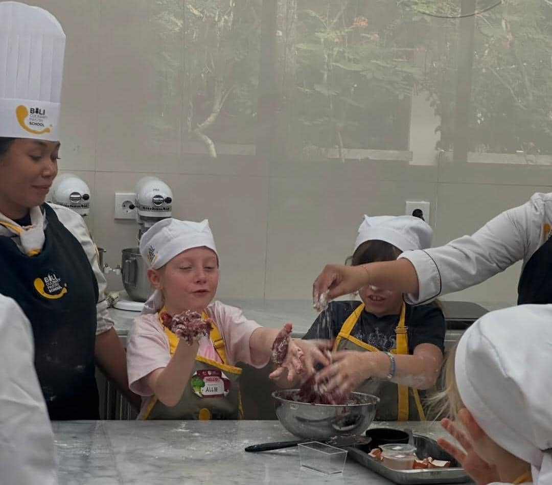 Anak-anak mengenakan celemek dan topi koki sedang belajar memasak bersama instruktur di kelas memasak di Bali.