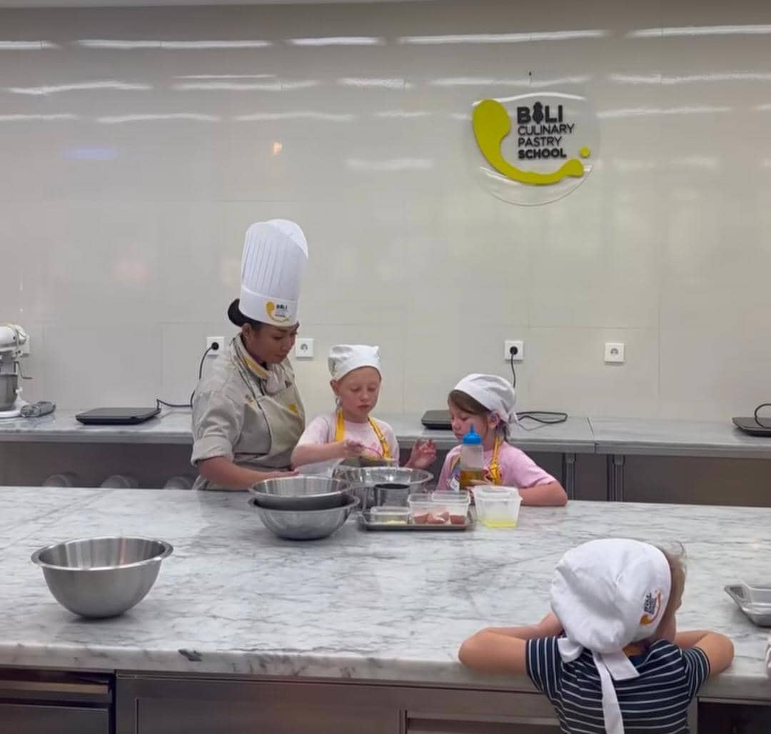 Seorang instruktur memasak membimbing dua anak kecil mengenakan celemek dan topi koki di dapur sekolah kuliner di Bali.