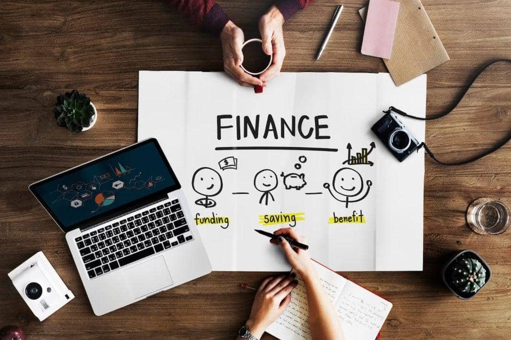 Tangan seseorang sedang menulis rencana keuangan bertuliskan finance, funding, saving, dan benefit di atas meja kerja.