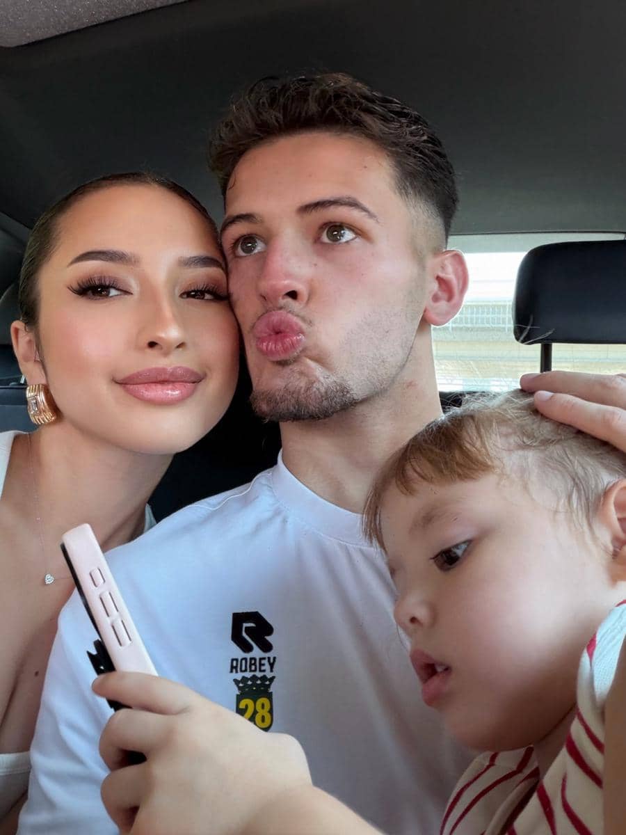 Seorang wanita, pria, dan anak kecil berfoto bersama di dalam mobil, tampak tersenyum dan memegang ponsel berwarna terang.