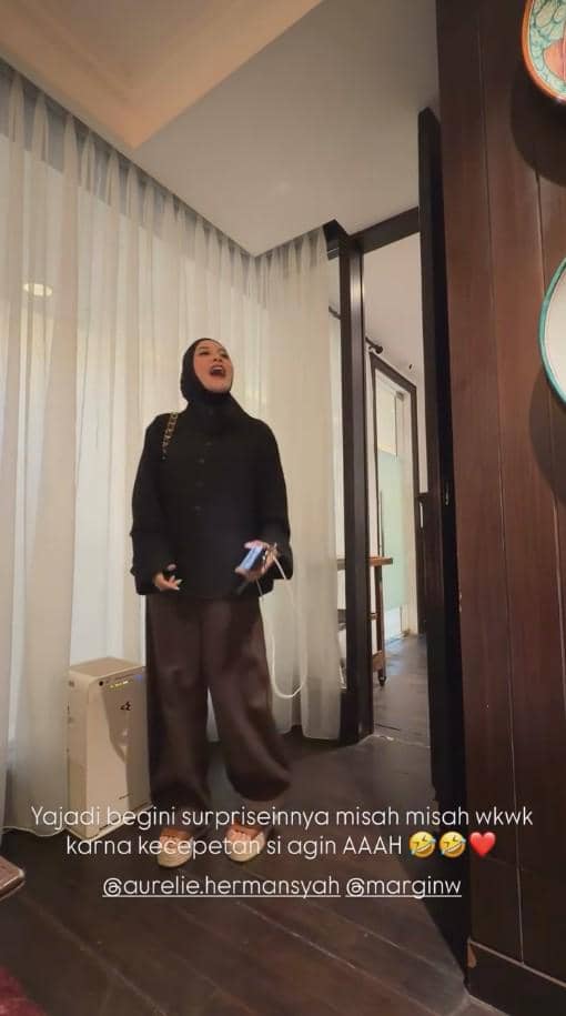 Seorang wanita berhijab hitam tertawa sambil memegang ponsel di dalam ruangan dengan tirai putih dan dinding kayu.