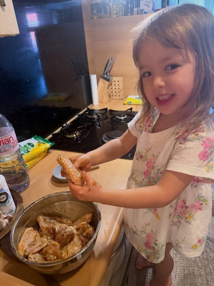 Seorang anak kecil mengenakan gaun bermotif bunga sedang memegang ayam mentah di dapur sambil tersenyum ke arah kamera.