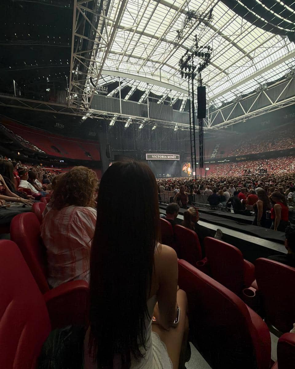Seorang perempuan berambut panjang duduk di kursi penonton stadion besar yang dipenuhi orang, menonton konser dengan panggung bertuliskan Victoria Monét.