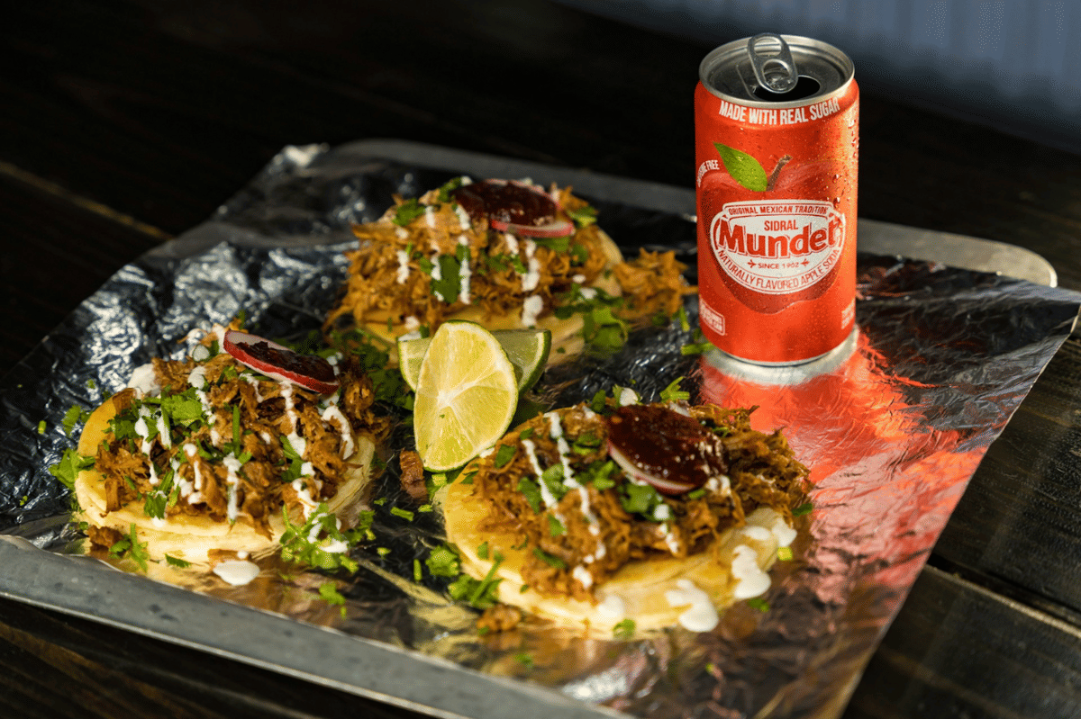 Tiga taco carnitas dengan daging suwir, saus, dan irisan jeruk nipis disajikan di atas aluminium foil bersama satu kaleng minuman Sidral Mundet.