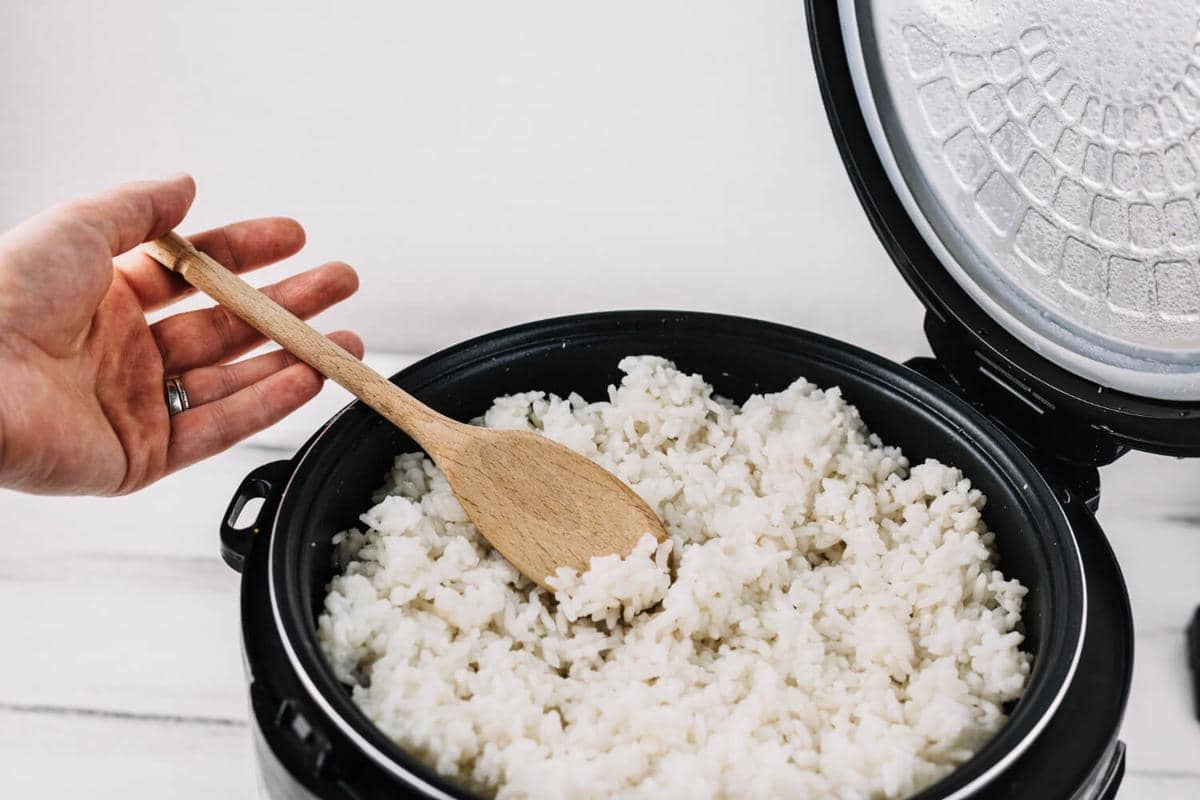 Ilustrasi semangkuk nasi putih di atas meja dengan rice cooker dan penghangat nasi sebagai peralatan dapur.