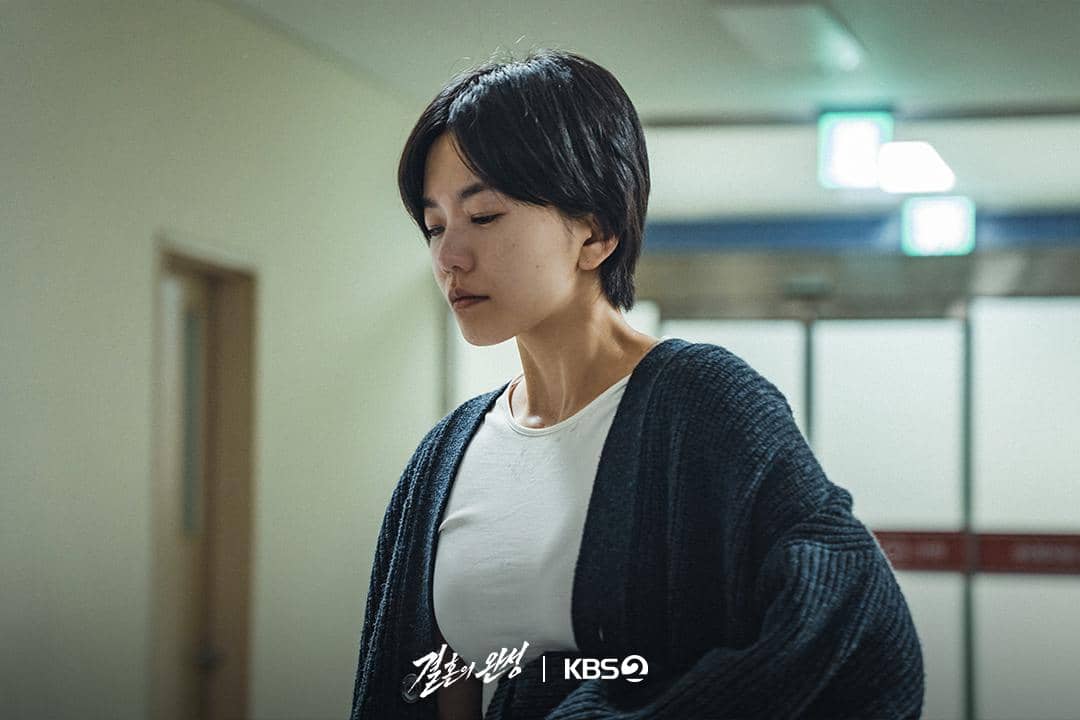 Seorang wanita berambut pendek mengenakan kaus putih dan kardigan gelap tampak menunduk di lorong gedung dalam cuplikan drama The Husband.