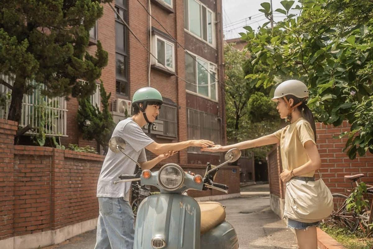 Dua remaja mengenakan helm saling menyentuh tangan di jalan sempit dengan latar bangunan bata merah dan sepeda motor biru di antara mereka.