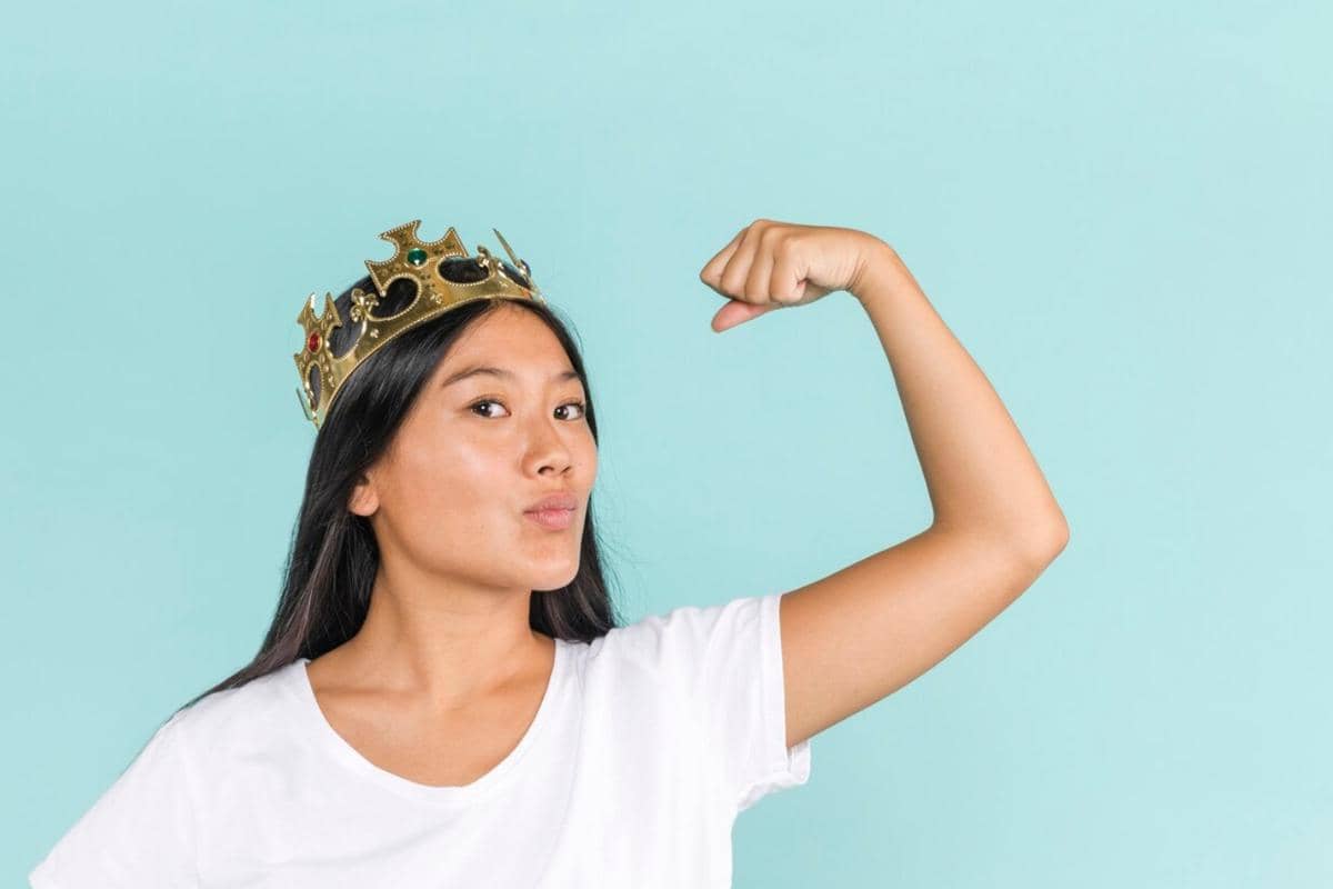 Seorang perempuan mengenakan mahkota emas sambil menunjukkan otot lengannya dengan ekspresi percaya diri di latar biru muda.