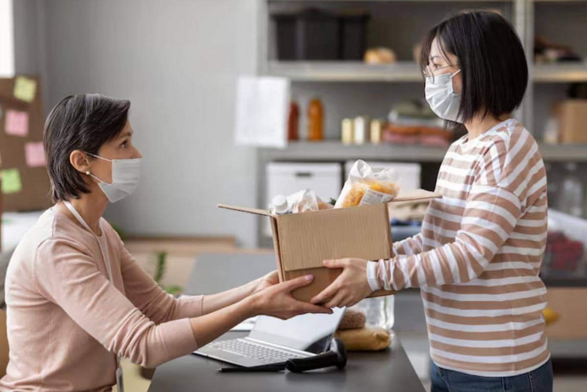 Dua perempuan mengenakan masker saling menyerahkan kotak berisi barang di meja dalam ruangan dengan rak penyimpanan di belakang.