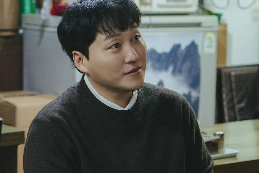 Seorang pria berambut pendek mengenakan sweater cokelat duduk di dalam ruangan dengan ekspresi serius dalam adegan drama The Husband.