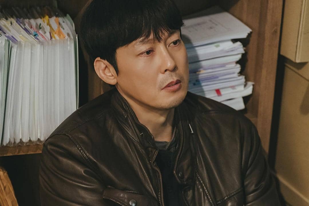 Seorang pria berjaket kulit cokelat duduk di depan rak berisi tumpukan berkas dalam adegan drama The Husband.