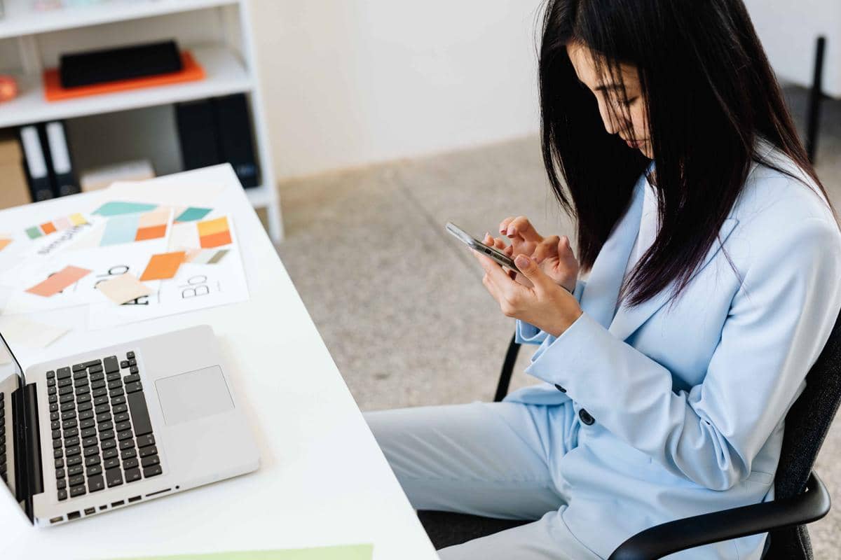 Seorang wanita mengenakan setelan biru muda duduk di meja kerja sambil menggulir ponsel di tangannya di dekat laptop terbuka.