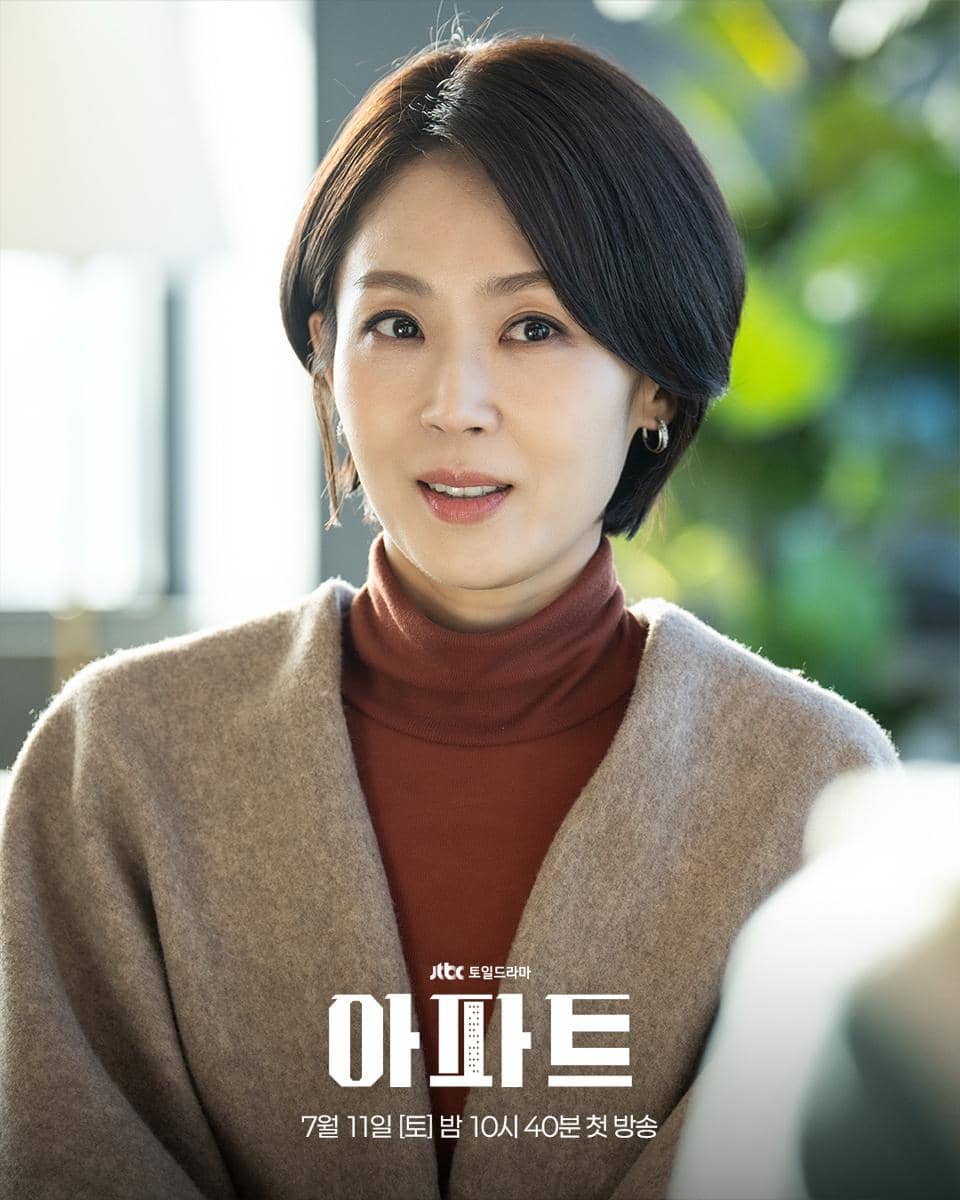 Seorang wanita berambut pendek mengenakan sweater cokelat muda dan turtleneck merah bata sedang tersenyum di adegan drama The Apartment Job.