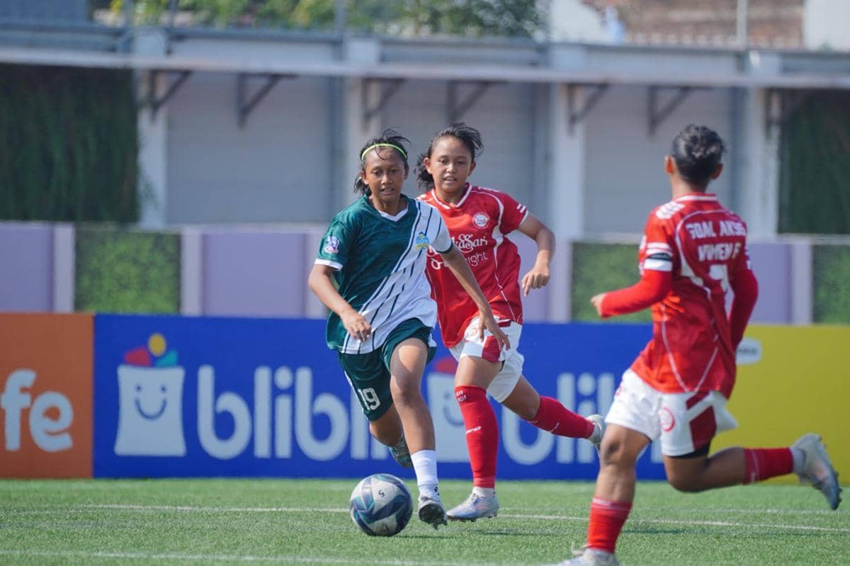 Pemain sepak bola wanita mengenakan seragam hijau menggiring bola di lapangan saat pertandingan melawan tim berseragam merah.