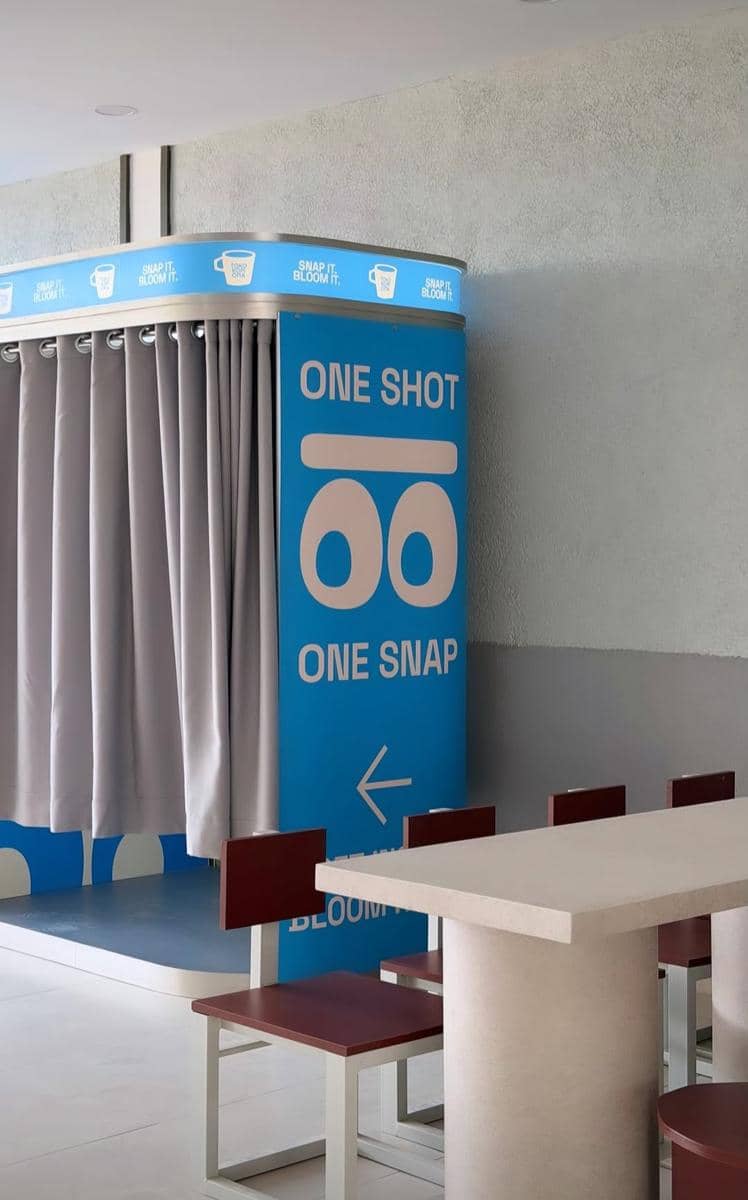 Sudut interior Toko Kopi Oma cabang Way Halim dengan bilik foto berwarna biru bertuliskan 'One Shot One Snap' dan meja kursi minimalis.