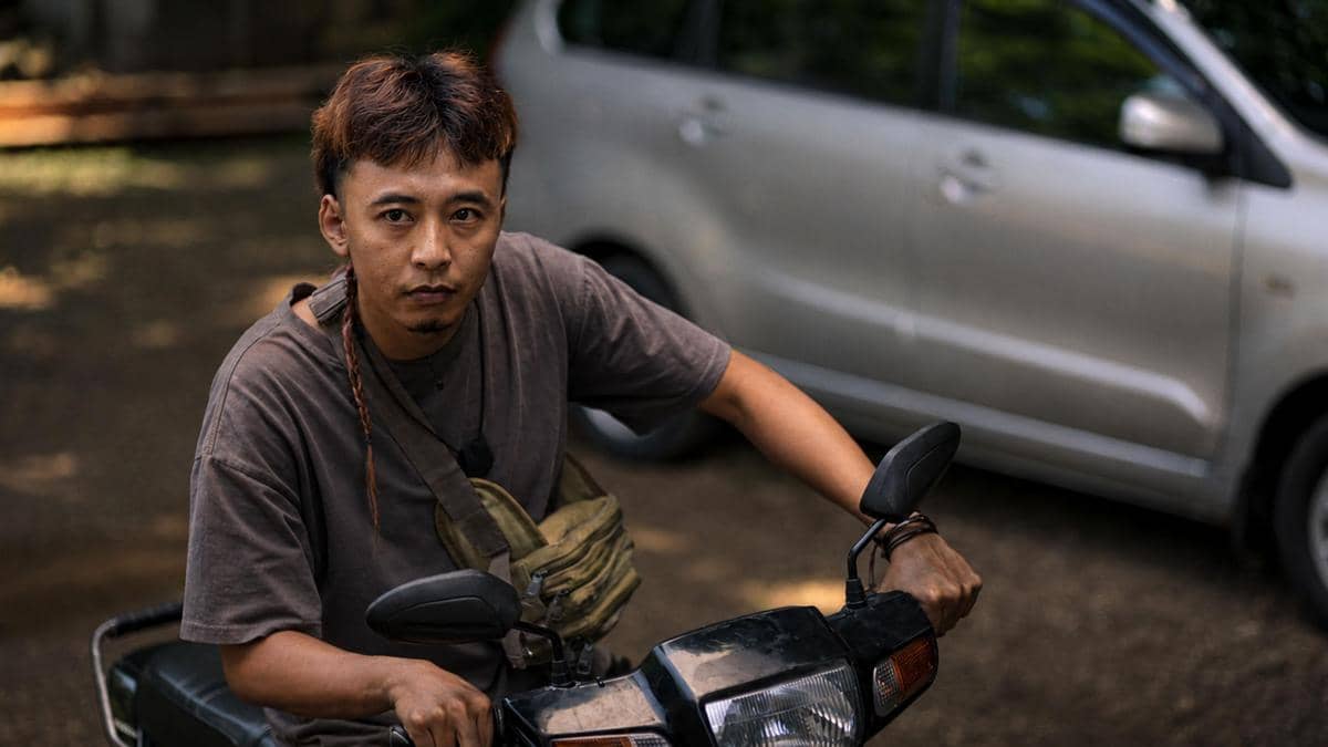 Seorang pria berambut pendek mengenakan kaus abu-abu duduk di atas motor dengan latar mobil perak di area luar ruangan.