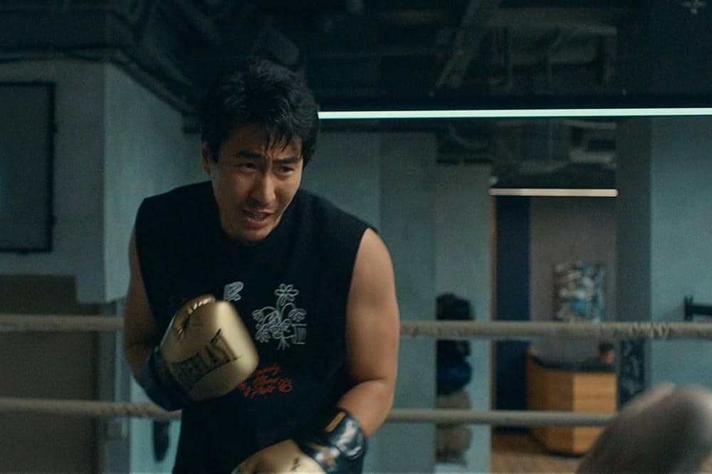 Seorang pria mengenakan sarung tinju emas dan kaus tanpa lengan hitam sedang berlatih tinju di dalam ring dengan latar gym modern.