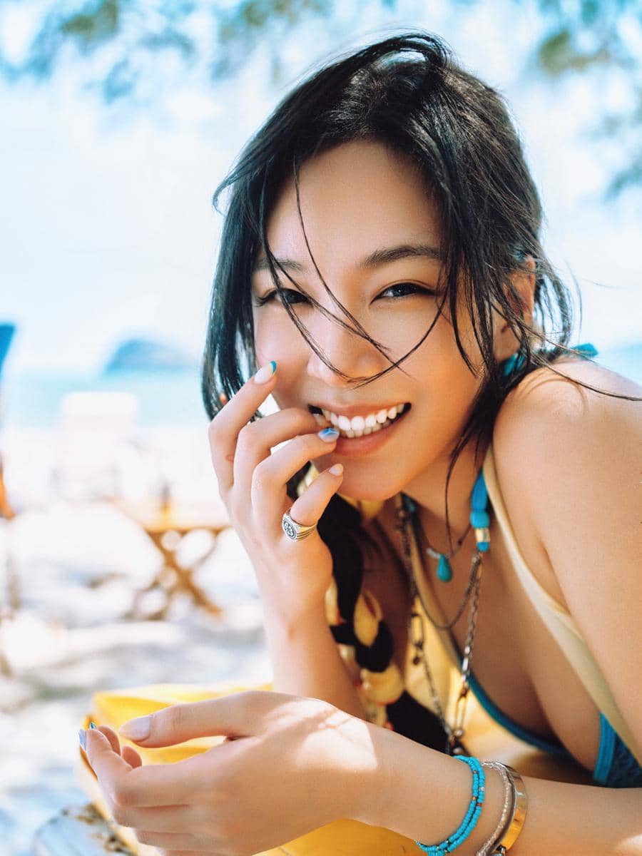 Seorang perempuan tersenyum cerah di pantai dengan latar laut biru, mengenakan aksesori berwarna turquoise dan pakaian musim panas.