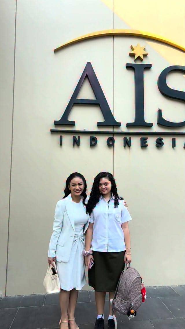 Seorang wanita berbusana putih berdiri bersama anak berseragam sekolah di depan gedung bertuliskan AIS Indonesia pada hari pertama sekolah.
