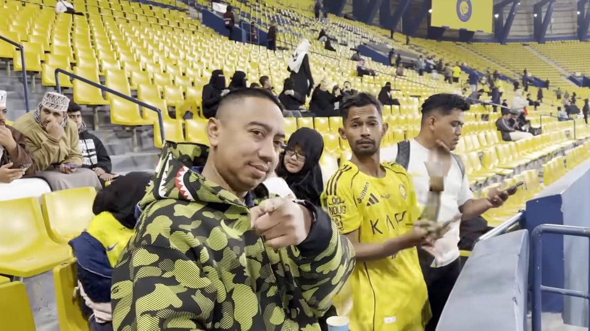 Dua pria berdiri di tribun Stadion Al-Awwal Arab Saudi dengan latar kursi kuning dan penonton mengenakan pakaian hitam di belakang mereka.