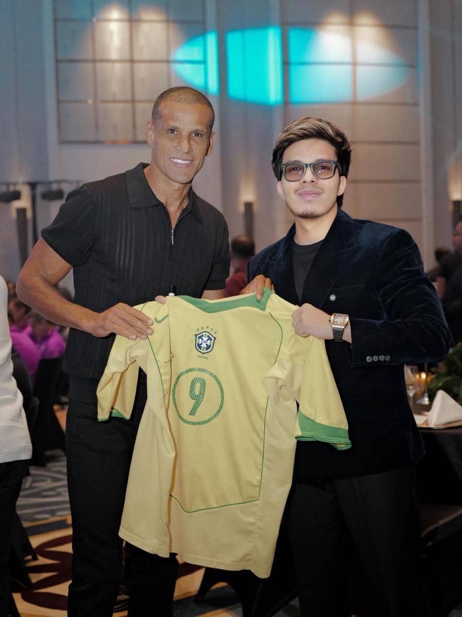 Dua pria berpose sambil memegang jersey tim nasional Brasil bernomor 9 di sebuah acara dalam ruangan dengan pencahayaan hangat.