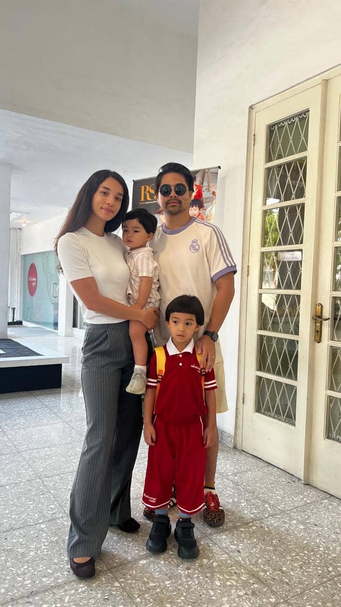 Seorang perempuan dan laki-laki berpose bersama dua anak kecil di depan pintu kaca, salah satu anak mengenakan seragam sekolah merah.
