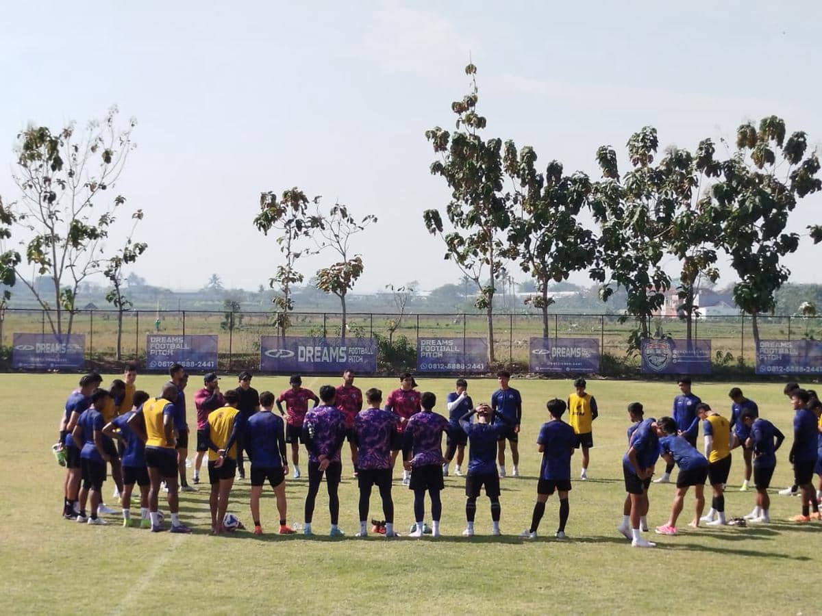 Para pemain Arema FC berkumpul di lapangan saat sesi latihan di bawah sinar matahari dengan latar pepohonan dan pagar.