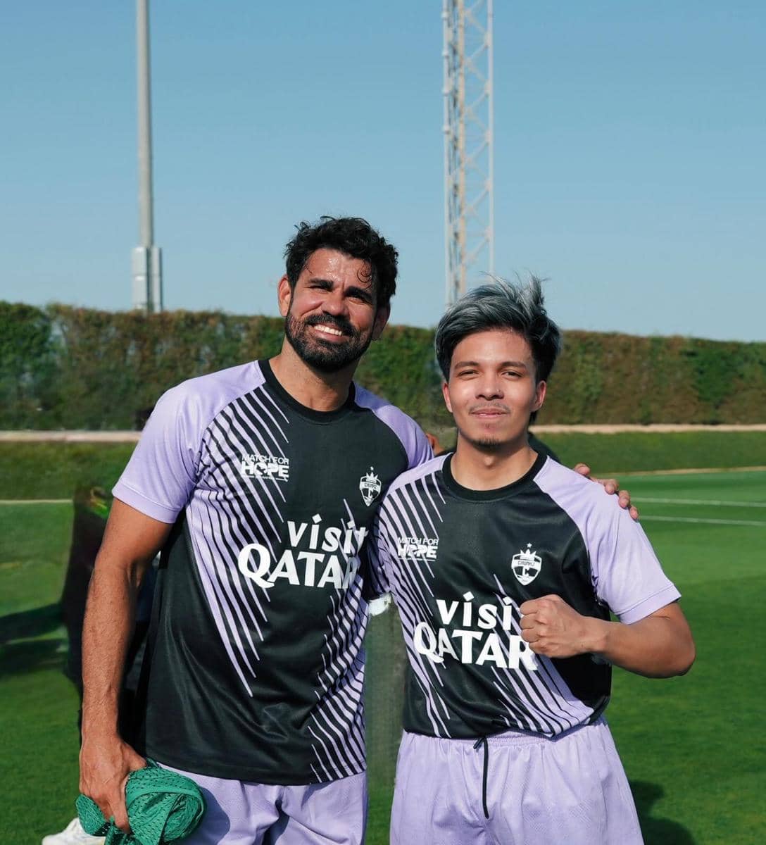 Dua pria mengenakan seragam sepak bola berwarna hitam dan ungu dengan tulisan 'Visit Qatar' berpose di lapangan hijau di bawah langit cerah.