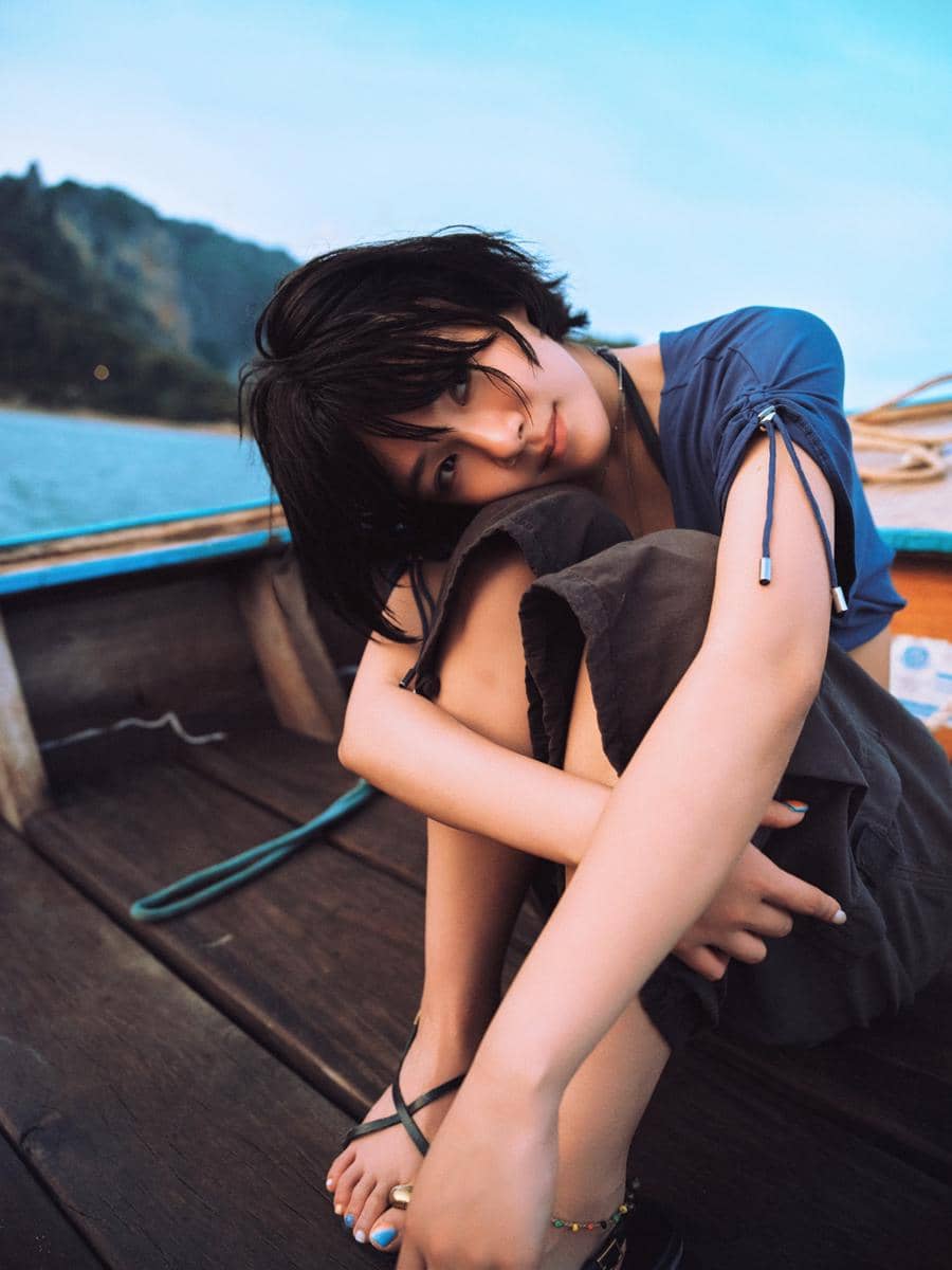 Seorang perempuan berambut pendek duduk di atas perahu kayu dengan latar laut dan pegunungan saat senja, mengenakan pakaian kasual.