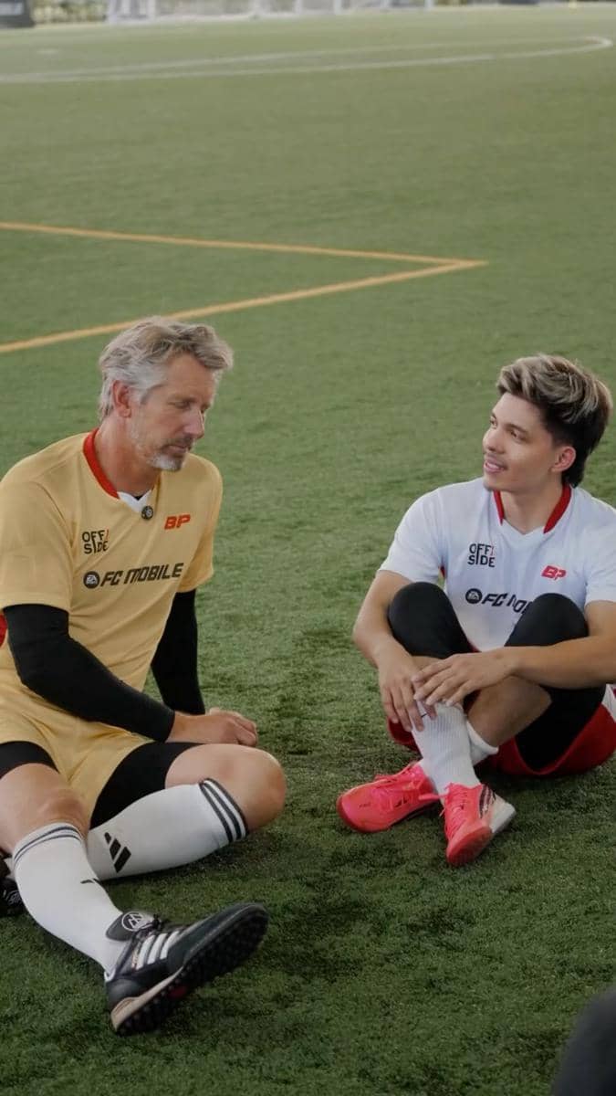 Dua pria mengenakan seragam sepak bola duduk di lapangan hijau sambil berbincang santai setelah bermain bola.