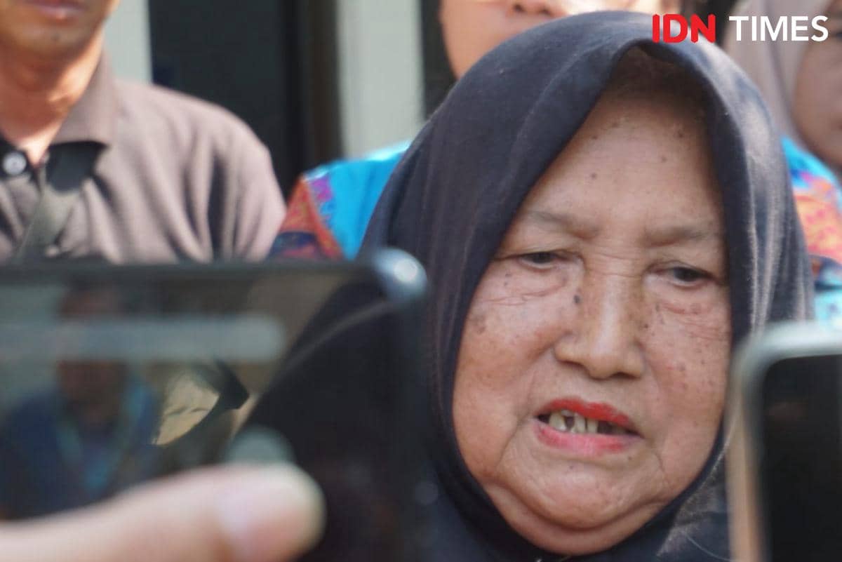 Seorang perempuan lanjut usia berhijab hitam berbicara di depan kamera dengan ekspresi serius, dikelilingi beberapa orang di sekitarnya.