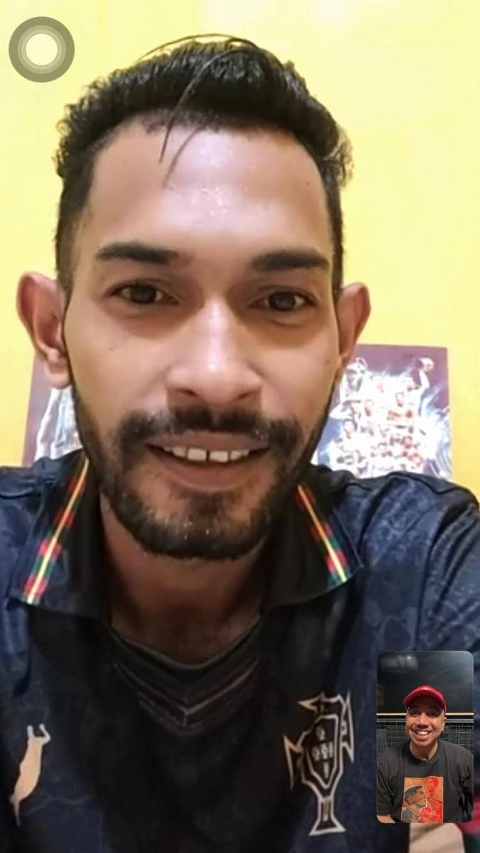 Seorang pria tersenyum saat melakukan video call dengan pria lain yang muncul di layar kecil di sudut bawah kanan.