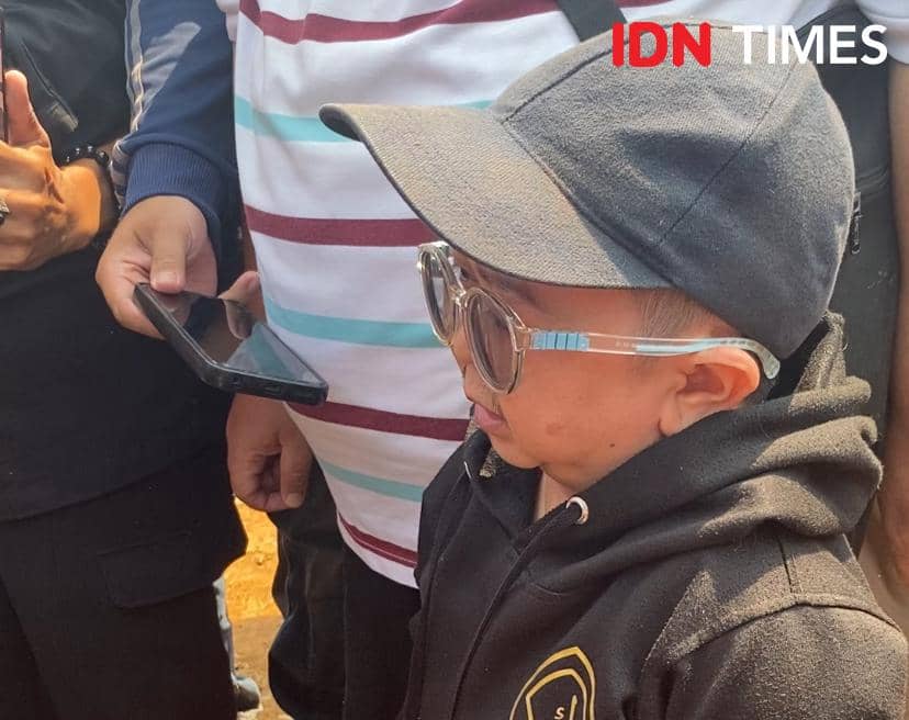 Seorang pria mengenakan jaket hitam, topi, dan kacamata besar berdiri di antara orang-orang saat prosesi pemakaman di TPU Tanah Kusir.