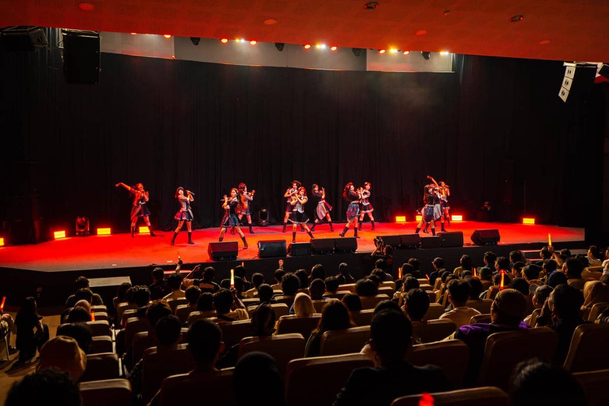 Penampilan grup idola di atas panggung dengan pencahayaan merah di depan penonton dalam tur teater sementara di Surabaya dan Yogyakarta.