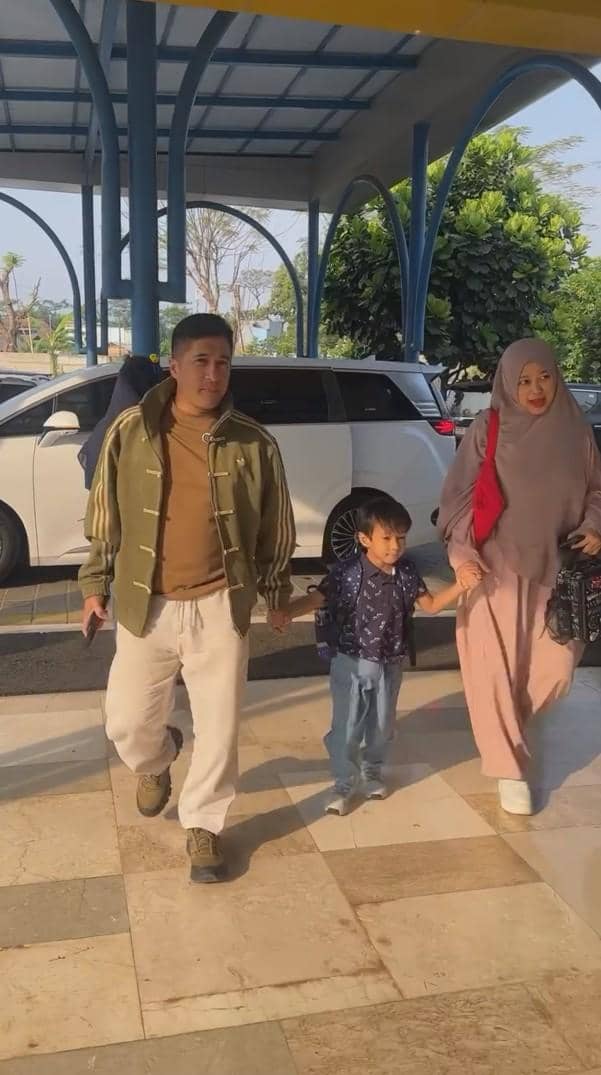 Seorang pria dan wanita berjalan bersama anak kecil menuju area sekolah pada pagi hari, tampak suasana cerah dan kendaraan di belakang mereka.